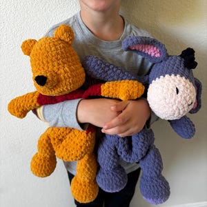 May include: A child holding two handmade crochet plush toys. One is yellow with a red shirt, resembling a bear. The other is a purple donkey with a pink inner ear and a white muzzle. Both toys are soft and cuddly.