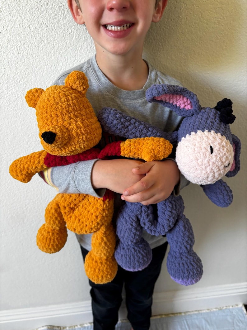 May include: A child holding two handmade crochet plush toys. One is yellow with a red shirt, resembling a bear. The other is a purple donkey with a pink inner ear and a white muzzle. Both toys are soft and cuddly.
