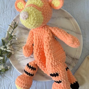 May include: A handmade orange and yellow crochet tiger plush toy. The tiger has black stripes on its legs and tail, with yellow accents on its ears and face. The plush is lying on a marble plate.