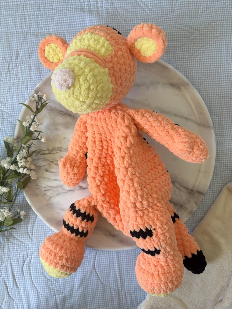 May include: A handmade orange and yellow crochet tiger plush toy. The tiger has black stripes on its legs and tail, with yellow accents on its ears and face. The plush is lying on a marble plate.