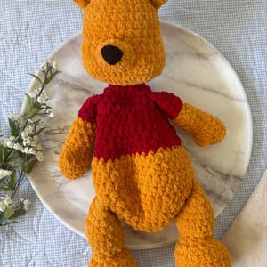 May include: A handmade crochet plush toy in the shape of a bear. The bear is primarily orange with a red shirt and a black nose. The toy is lying on a white marble plate.