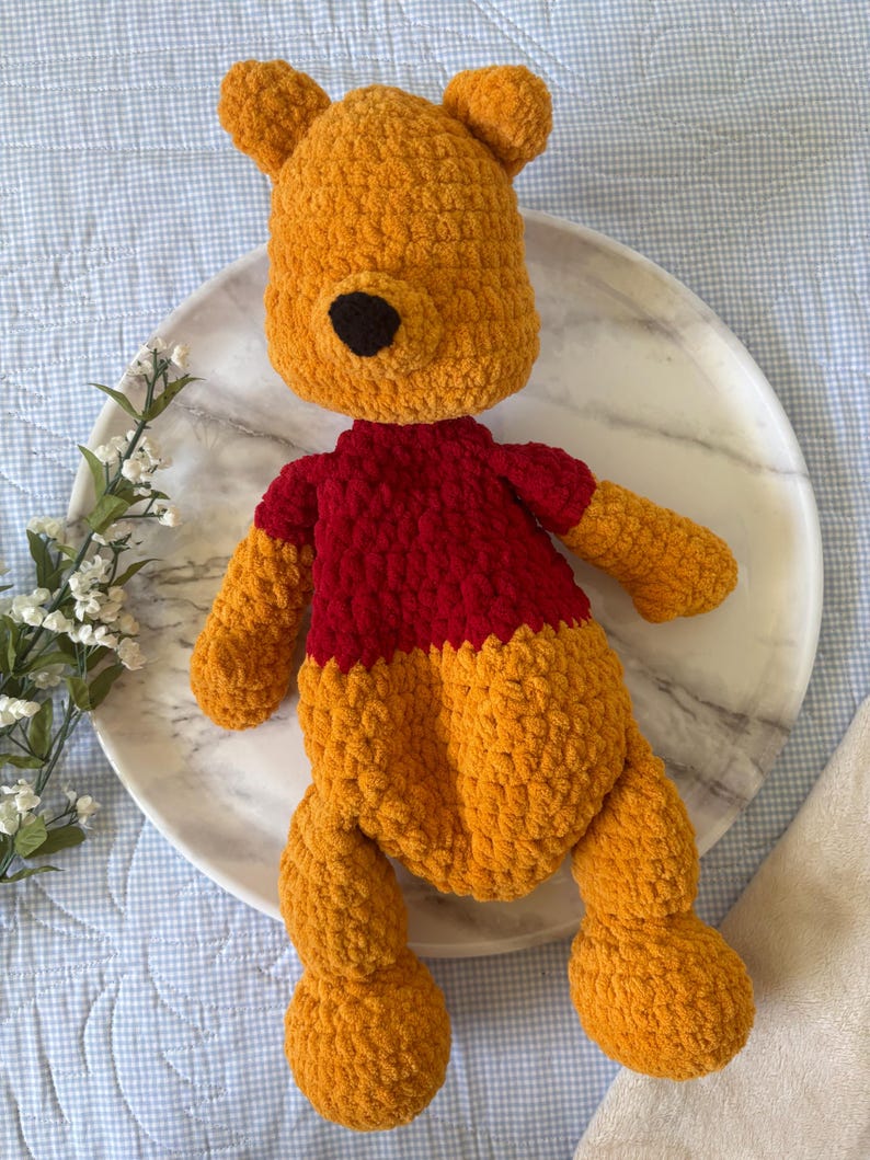May include: A handmade crochet plush toy in the shape of a bear. The bear is primarily orange with a red shirt and a black nose. The toy is lying on a white marble plate.