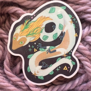 Könnte beinhalten: Ein weißer und goldener Drachenaufkleber mit grünen Blättern und goldenen Sternen auf schwarzem Hintergrund. Der Drache ist zu einem Kreis zusammengerollt, mit dem Kopf nach links gerichtet.