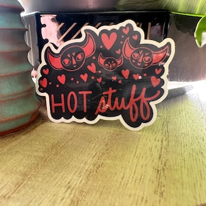 May include: A black and red sticker with three cartoon cat faces and the text "HOT stuff" in red. The sticker is surrounded by red hearts.