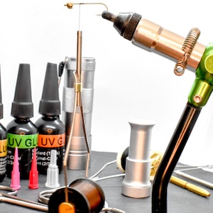May include: A fly tying vise with various tools and materials. The image shows bottles of UV glue, scissors, thread, and other accessories. The vise is black, green, and copper-colored, with a hook attached. The glue bottles have black caps and colorful labels.