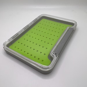 May include: A rectangular fly fishing box with a clear lid and a gray frame. The interior features a bright green foam with numerous small, triangular slots for securing flies. The box is designed to protect and organize fishing flies.