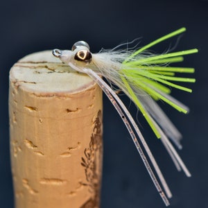 May include: A white and green fly fishing lure with a silver bead head. The lure is tied on a cork.