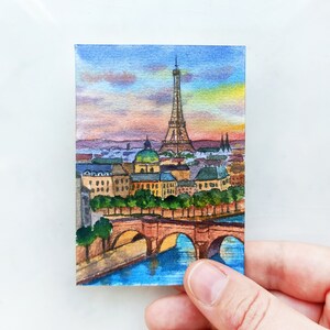 May include: Watercolor painting of the Eiffel Tower in Paris, France. The painting depicts the tower with a bridge in the foreground and a cityscape in the background. The sky is a soft blue with pink and orange clouds.