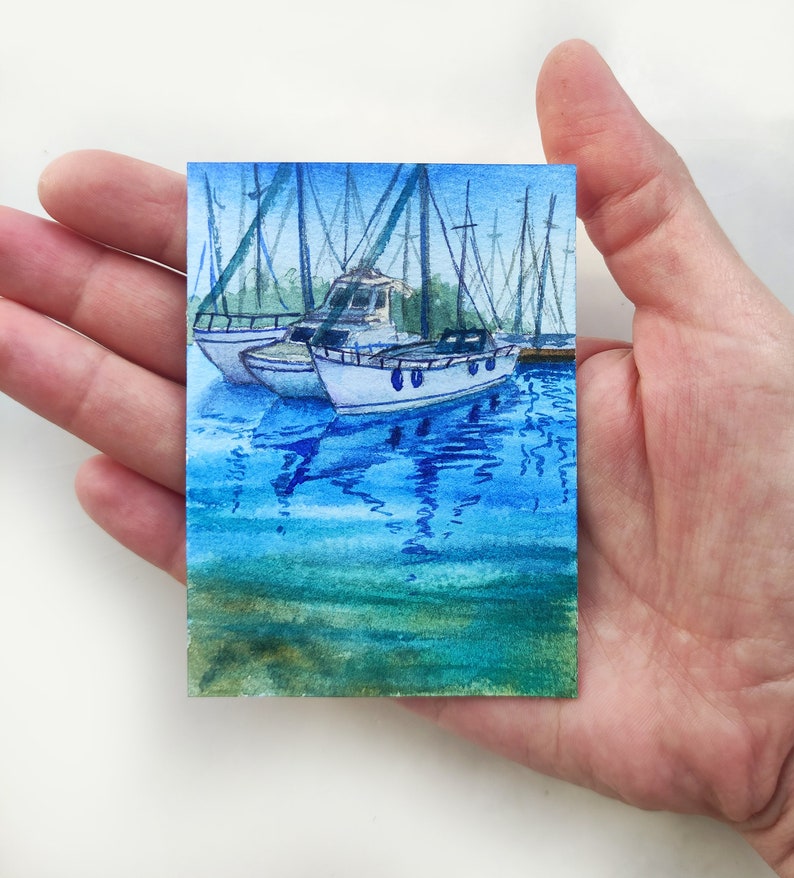 Puede incluir: Pintura acuarela de un puerto deportivo con varios veleros atracados. Los barcos son blancos con detalles azules y se reflejan en el agua azul. El agua est&aacute; pintada en tonos azules y verdes.
