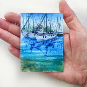 Puede incluir: Pintura acuarela de una escena de puerto con varios veleros atracados. Los barcos son blancos con detalles azules y se reflejan en el agua azul. El agua es de color azul claro con ondas azules m&aacute;s oscuras. El cielo es de color azul claro.