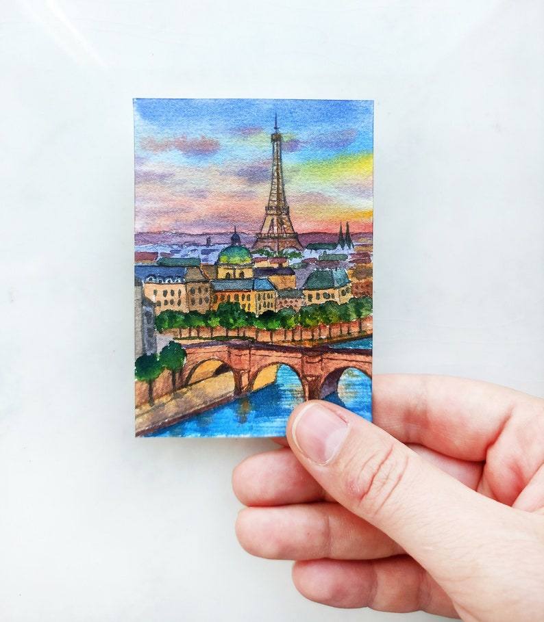 May include: Watercolor painting of the Eiffel Tower in Paris, France. The painting depicts the tower with a bridge in the foreground and a cityscape in the background. The sky is a soft blue with pink and orange clouds.