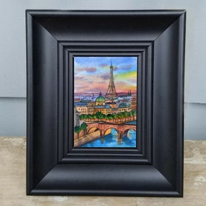 May include: A framed watercolor painting of the Eiffel Tower in Paris, France. The painting depicts the tower in the distance with a bridge and river in the foreground. The sky is a vibrant sunset with pink, orange, and purple hues.
