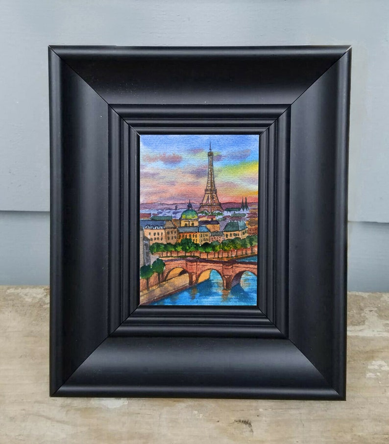 May include: A framed watercolor painting of the Eiffel Tower in Paris, France. The painting depicts the tower in the distance with a bridge and river in the foreground. The sky is a vibrant sunset with pink, orange, and purple hues.