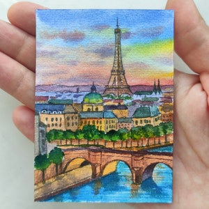 May include: Watercolor painting of the Eiffel Tower in Paris, France. The painting depicts the iconic tower with a bridge in the foreground and a cityscape in the background. The sky is a vibrant sunset with shades of orange, pink, and purple.