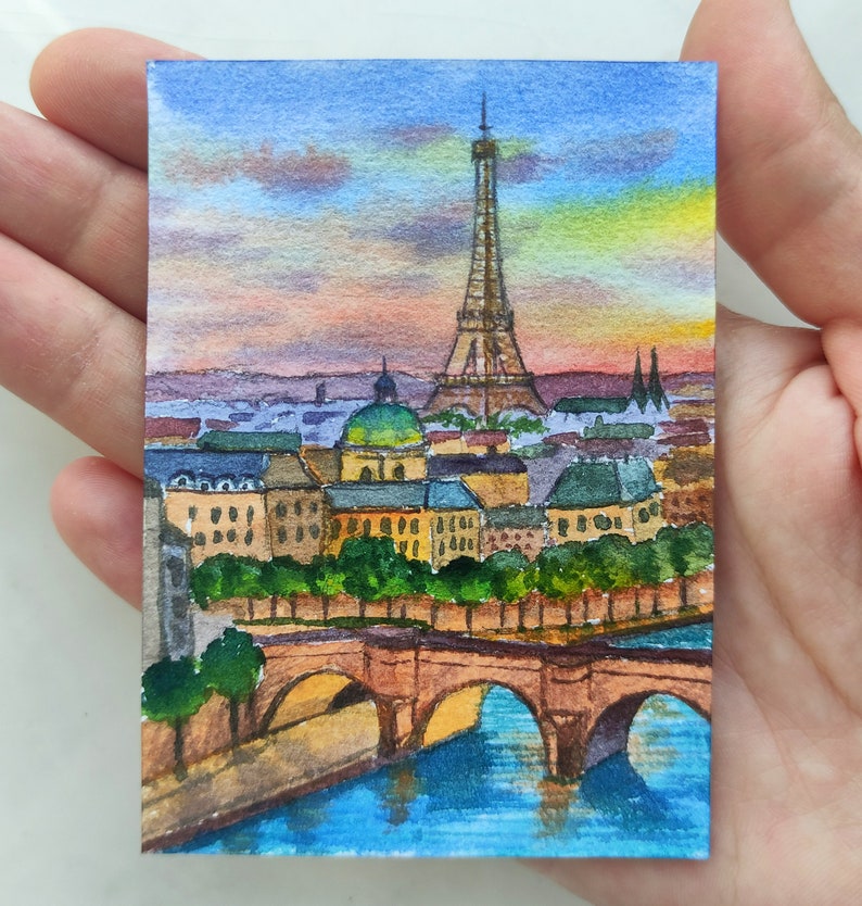 May include: Watercolor painting of the Eiffel Tower in Paris, France. The painting depicts the iconic tower with a bridge in the foreground and a cityscape in the background. The sky is a vibrant sunset with shades of orange, pink, and purple.