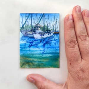 Puede incluir: Pintura acuarela de dos veleros blancos atracados en un puerto con agua azul y reflejos. La pintura est&aacute; en una tarjeta peque&ntilde;a.