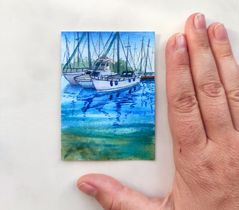 Puede incluir: Pintura acuarela de dos veleros blancos atracados en un puerto con agua azul y reflejos. La pintura est&aacute; en una tarjeta peque&ntilde;a.
