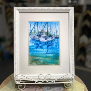 Puede incluir: Una pintura de acuarela enmarcada de tres veleros atracados en un puerto. Los barcos son blancos con detalles azules y est&aacute;n rodeados de agua azul con reflejos. La pintura est&aacute; enmarcada en un marco blanco con un borde decorativo.