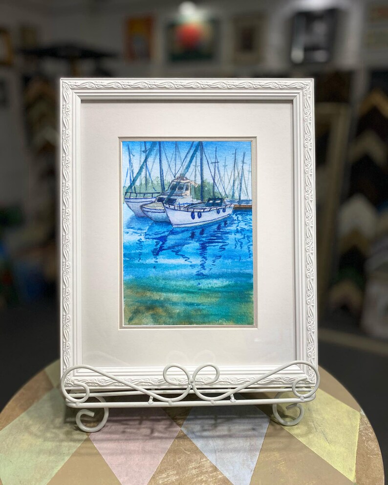 Puede incluir: Una pintura de acuarela enmarcada de tres veleros atracados en un puerto. Los barcos son blancos con detalles azules y est&aacute;n rodeados de agua azul con reflejos. La pintura est&aacute; enmarcada en un marco blanco con un borde decorativo.