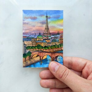 May include: Watercolor painting of the Eiffel Tower in Paris, France. The painting depicts the tower in the distance with a bridge and buildings in the foreground. The sky is a vibrant blue with pink and orange clouds.