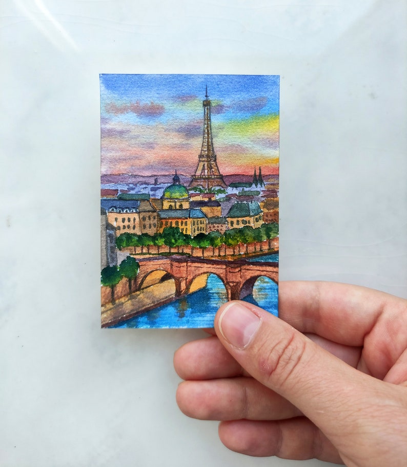 May include: Watercolor painting of the Eiffel Tower in Paris, France. The painting depicts the tower in the distance with a bridge and buildings in the foreground. The sky is a vibrant blue with pink and orange clouds.