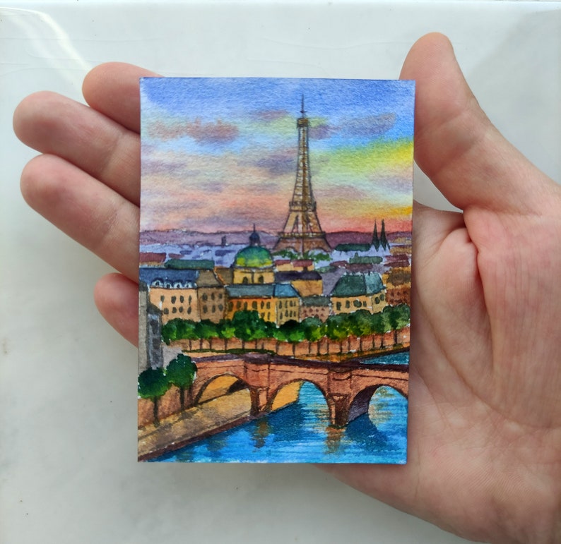 May include: Watercolor painting of the Eiffel Tower in Paris, France. The painting depicts the tower with a bridge in the foreground and a sunset sky in the background.