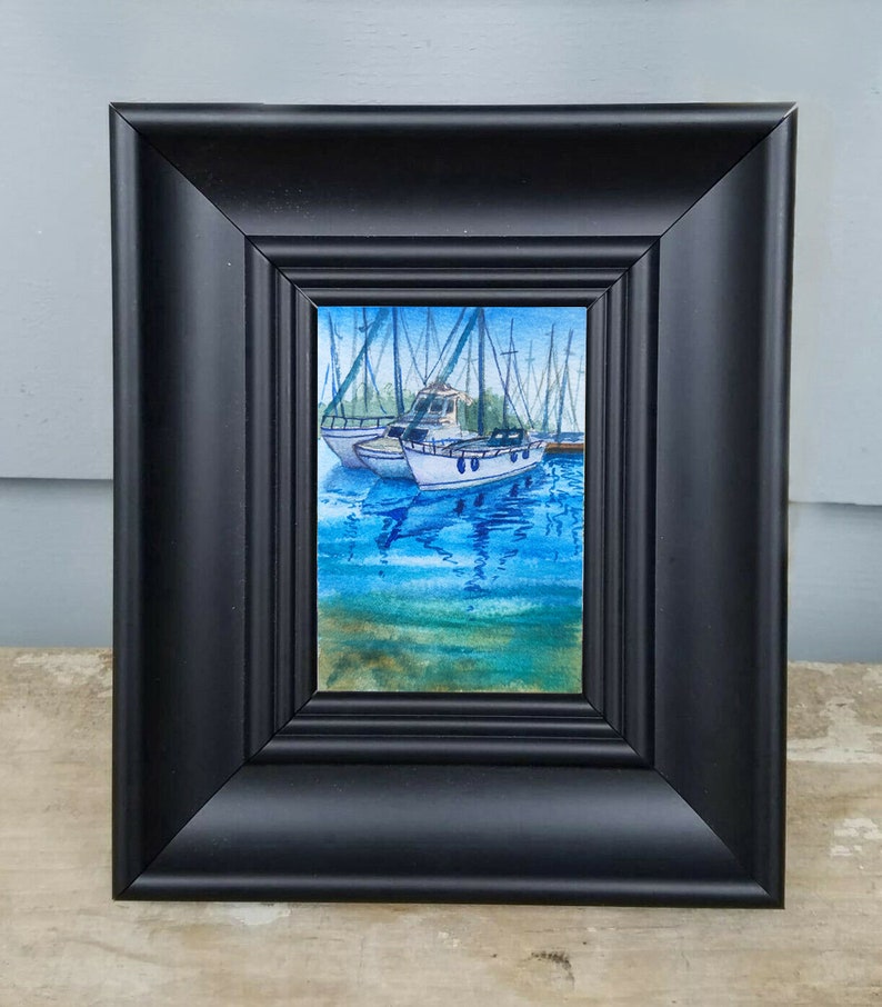 Puede incluir: Una pintura de acuarela enmarcada de dos veleros blancos atracados en un puerto deportivo. Los barcos est&aacute;n rodeados de agua azul con reflejos de los barcos y el cielo. La pintura est&aacute; enmarcada en un marco negro.