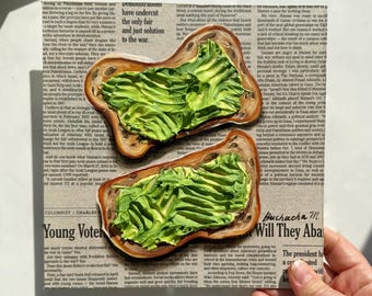 May include: Two slices of toasted bread topped with a generous layer of bright green avocado spread. The avocado is spread in a thick, textured layer, resembling a painting. The bread is brown and slightly toasted. The image is taken from a close-up perspective, with the avocado spread taking up most of the frame. The background is a newspaper with text that is partially visible.