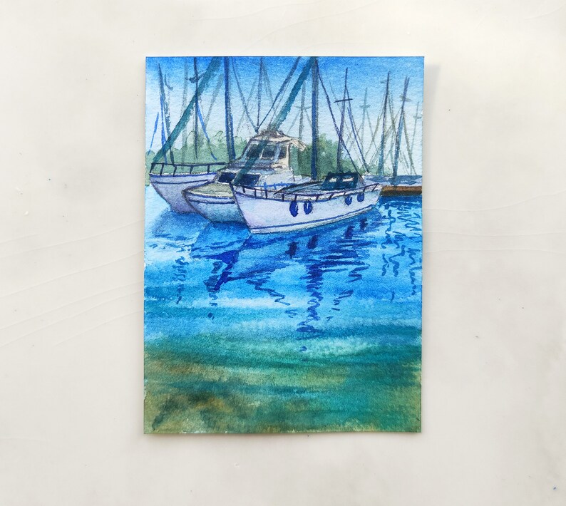 Puede incluir: Pintura acuarela de un puerto deportivo con varios veleros atracados. Los barcos son blancos con detalles azules y se reflejan en el agua azul. El agua es de color azul claro con ondas azules m&aacute;s oscuras. El cielo es de color azul claro.