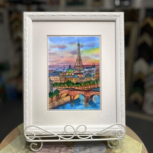 May include: A framed watercolor painting of the Eiffel Tower in Paris, France. The painting depicts the iconic landmark with a bridge in the foreground and a cityscape in the background. The sky is a vibrant sunset with shades of pink, orange, and purple.