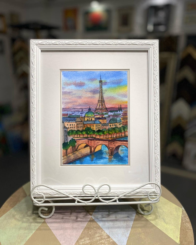 May include: A framed watercolor painting of the Eiffel Tower in Paris, France. The painting depicts the iconic landmark with a bridge in the foreground and a cityscape in the background. The sky is a vibrant sunset with shades of pink, orange, and purple.