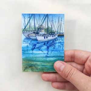 Puede incluir: Pintura acuarela de un peque&ntilde;o barco atracado en un puerto con otros barcos en el fondo. El agua es de color azul claro con reflejos de los barcos. El cielo es de color azul claro.