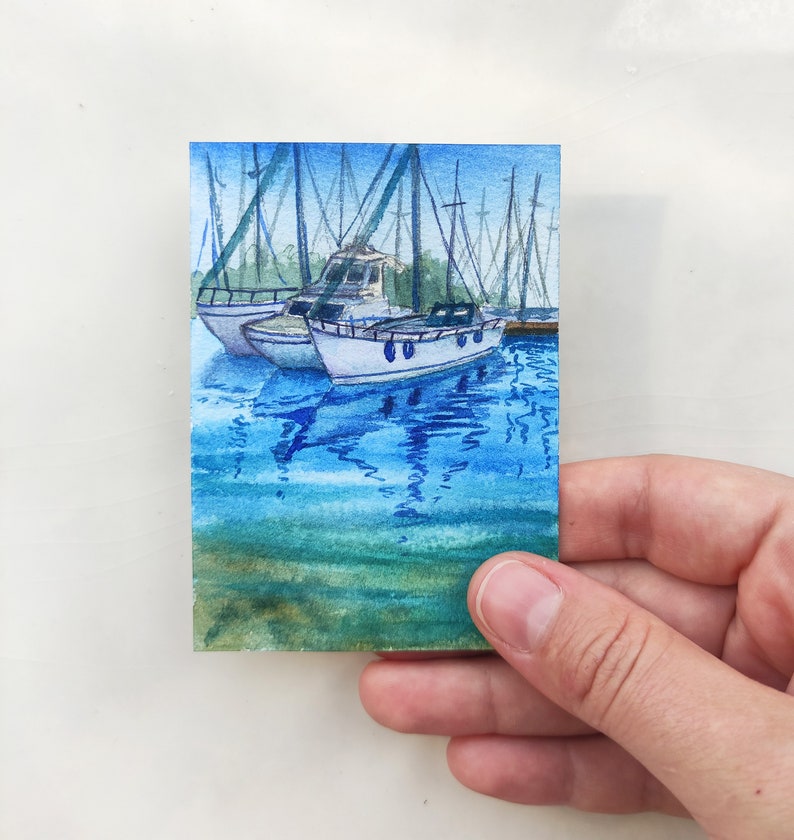 Puede incluir: Pintura acuarela de un peque&ntilde;o barco atracado en un puerto con otros barcos en el fondo. El agua es de color azul claro con reflejos de los barcos. El cielo es de color azul claro.