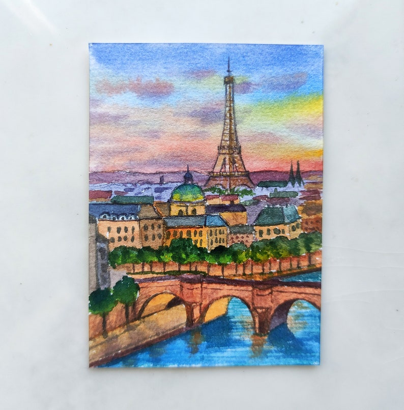 May include: Watercolor painting of the Eiffel Tower in Paris, France. The painting depicts the tower in the distance with a bridge and river in the foreground. The sky is a soft pink and orange.