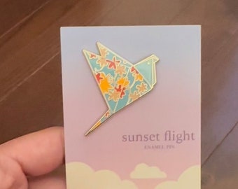 Könnte beinhalten: Eine blaue und goldene Origami-Vogel-Emaille-Anstecknadel mit einem Blumenmuster. Die Anstecknadel befindet sich auf einer Karte mit rosa Hintergrund und dem Text "sunset flight enamel pin".