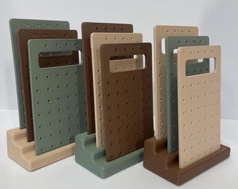 May include: A collection of earring display stands in various earth-tone colors, including beige, brown, and green. Each stand features a rectangular design with a handle and multiple holes for displaying earrings. The stands are arranged in a group.