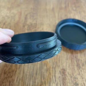 May include: Black plastic container with a diamond pattern on the base and a matching lid. The container has a textured surface and a circular opening. The lid is slightly ajar, revealing the interior.