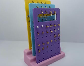 May include: A multi-colored earring display stand with three vertical panels in yellow, blue, and purple, each with multiple holes for earrings. The stand is on a pink base, showcasing various gold-colored earrings.