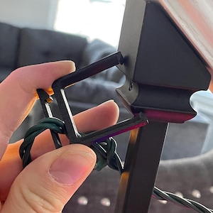 May include: A black plastic clip holding green Christmas lights. The clip is attached to a black metal post. The lights are wrapped around the clip. The background is blurred.