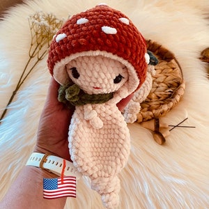 Op de afbeelding: Een handgemaakte gehaakte paddenstoelpop met een rode kap met witte stippen. De pop heeft een crèmekleurig lichaam, een groene sjaal en zwarte ogen. De pop is ongeveer 15 cm hoog.