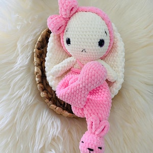 May include: A handmade, plush bunny doll with a white body, pink accents, and a bow. The bunny is seated in a woven basket on a fluffy, white surface. The bunny's ears, bow, and dress are a soft pink color.