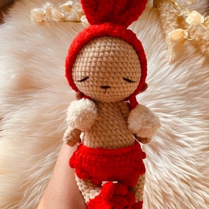 May include: A handmade crochet doll in the shape of a bunny. The doll has a red hood with heart-shaped ears, a red skirt, and white pom-poms. The body is beige, and the eyes are closed. A unique, handcrafted toy.