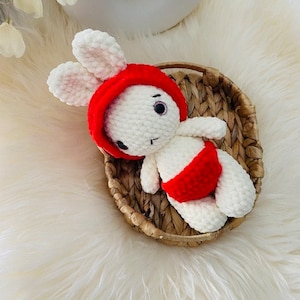 May include: A handmade white crochet bunny wearing a red hat and red diaper. The bunny is lying in a small woven basket on a white fluffy surface. The bunny has black eyes and a stitched mouth.