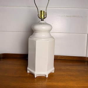 May include: A white ceramic table lamp with a brass-colored neck and handle. The lamp has a geometric, octagonal base and a rounded top. The lamp is approximately 12 inches tall.