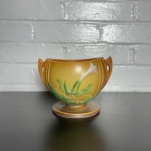 May include: A brown and green glazed ceramic planter with a white flower design and two handles. The planter has a pedestal base.