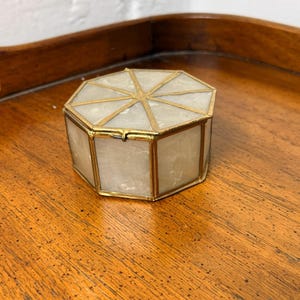 May include: An octagonal trinket box with a gold-colored metal frame and a translucent, off-white material for the sides and top. The box is resting on a wooden surface.