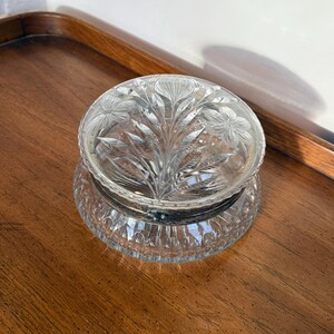 May include: A clear, round crystal trinket box with a floral design on the lid. The lid is domed and sits atop a base with a decorative, ridged pattern. The box is displayed on a wooden surface.