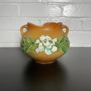 May include: A brown ceramic planter with a white and green floral design. The planter has two handles and a rounded shape.