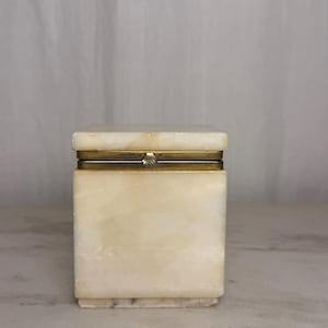 May include: A rectangular box crafted from smooth, cream-colored stone with natural veining. The box features a hinged lid with a metallic gold-toned rim and a small, decorative shell-shaped clasp centered on the front edge of the lid.