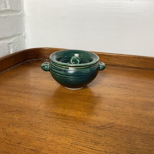 May include: A small green ceramic bowl with two handles. The bowl has a striped pattern and is sitting on a wooden surface.